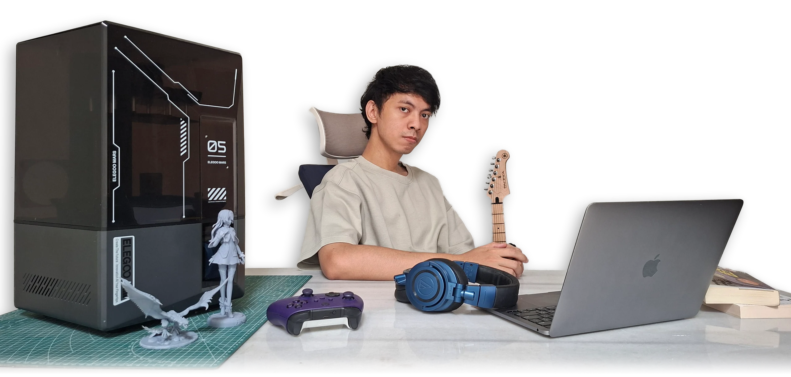 Wahyu at his desk with a resin printer, prints, a controller, headphones, a guitar and a laptop